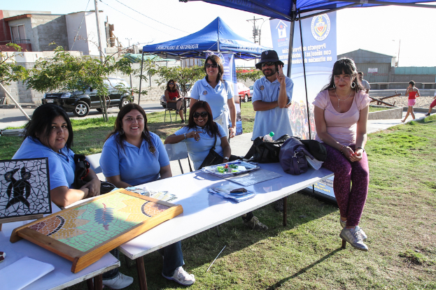 Eine Gruppe von Menschen sitzt an einem Tisch mit Spielen, Zelten und blauen Bannern im Hintergrund, während andere auf dem Rasen stehen, ein Fahrzeug, Bäume, ein Haus und der Himmel sichtbar sind.
