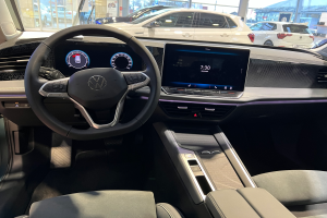 Das Innere eines Volkswagen e-Golf mit einem Lenkrad, Armaturenbrett, Bildschirmen, Knöpfen und Sitzen, mit einer Sicht auf andere Autos, Gebäude, Bäume, Pfosten und den Himmel durch die Glastüren.