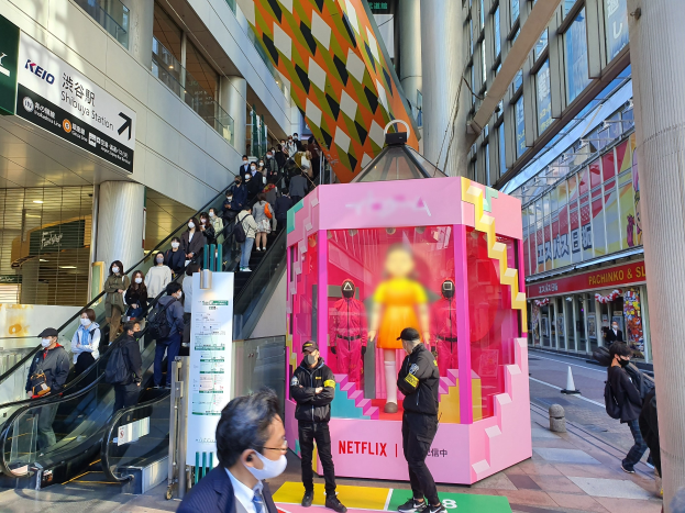 Eine Gruppe von Menschen steht vor einem gläsernen Gebäude mit Säulen und Texttafeln, einige tragen Masken, während eine Rolltreppe mit Menschen zu sehen ist; ein pinker Kasten in der Mitte zeigt Schaufensterpuppen und trägt den Text "Netflix's neuer Netflix-Stand in Tokyo."