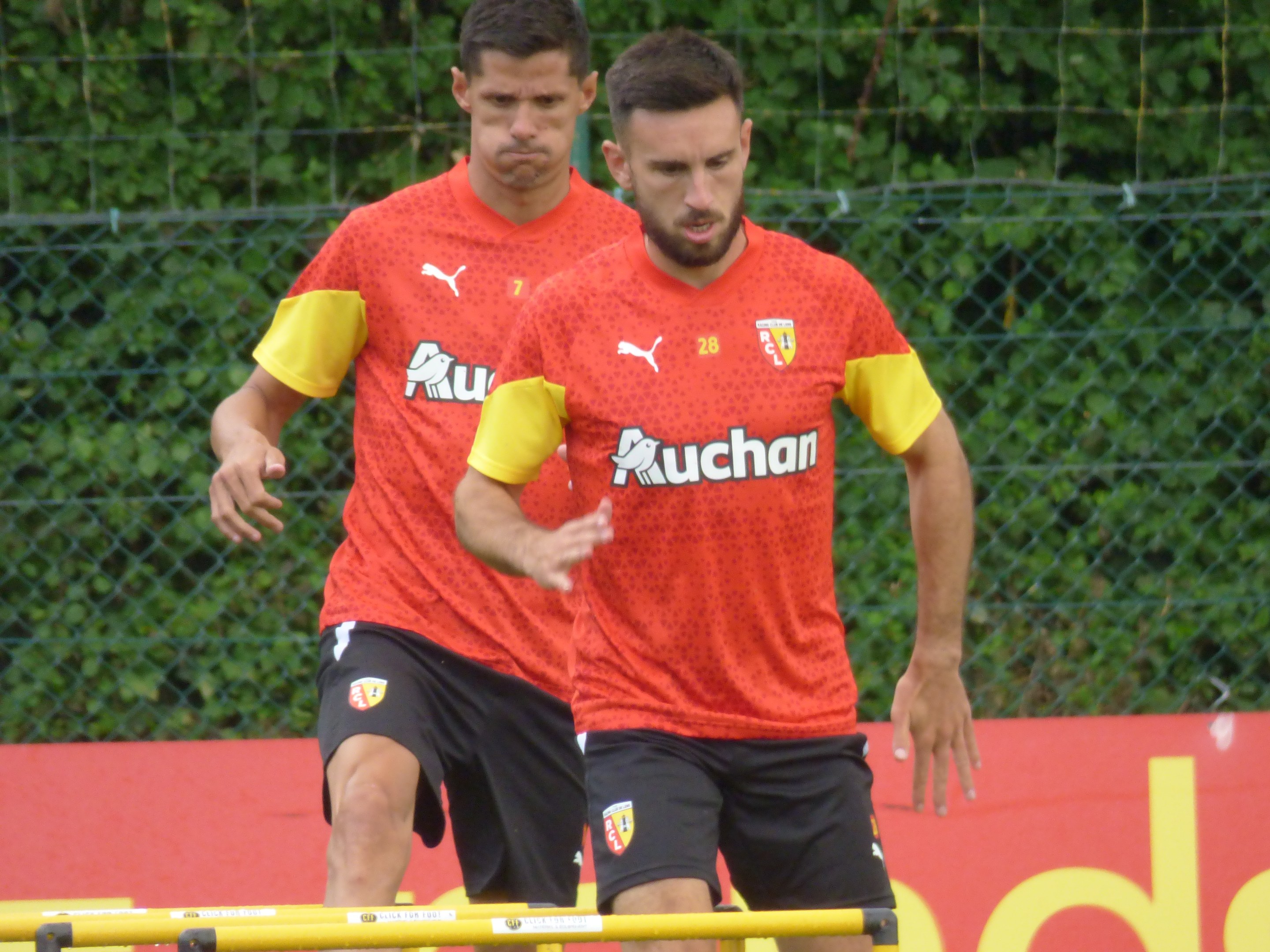 Two men in red shirts and black shorts playing soccer on a fenced field with trees in the background, with a red board displaying "Borussia Dortmund" in the distance.