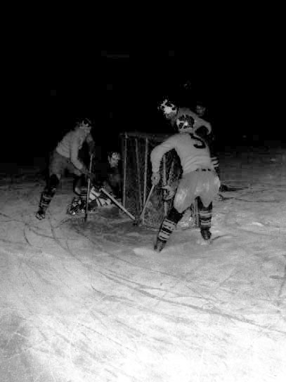 Schwarzes und weißes Foto von Männern, die bei Nacht auf dem Schnee Hockey spielen, tragen Helme und halten Stöcke.