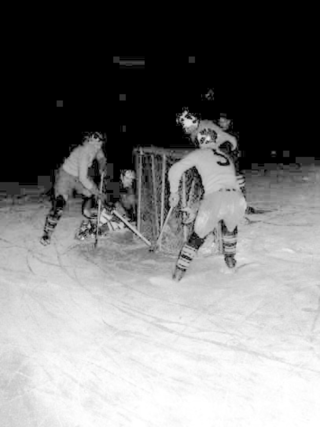 Schwarzes und weißes Foto von Männern, die bei Nacht auf dem Schnee Hockey spielen, tragen Helme und halten Stöcke.