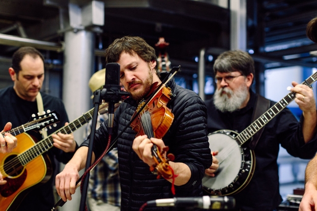 Eine Gruppe von Musikern in schwarzer Kleidung, die verschiedene Instrumente spielen, mit einem Geiger vor einem Mikrofon und einem Gebäude im Hintergrund.