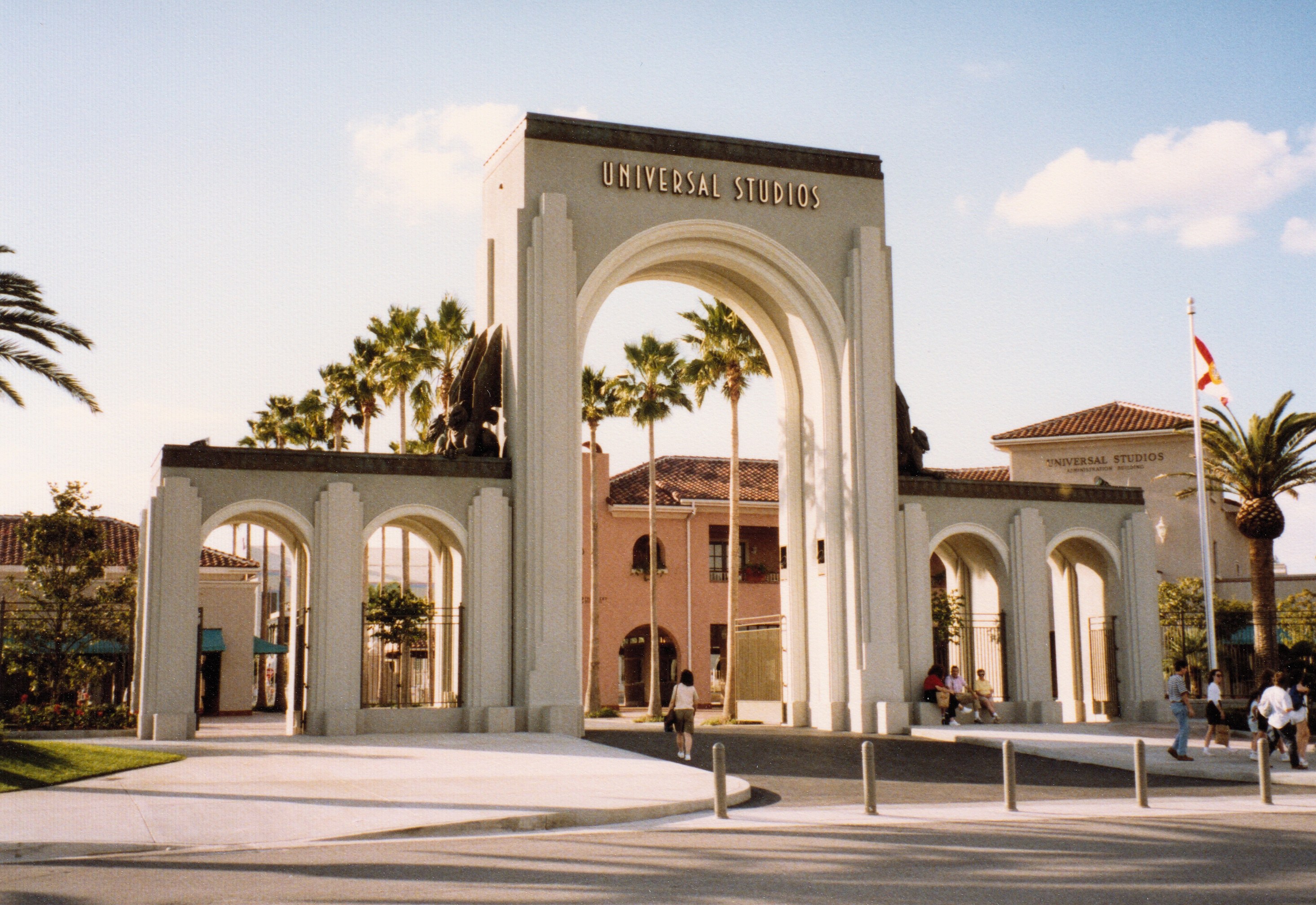 Eingang zu Universal Studios Hollywood mit einem Bogen, einer Statue, Menschen, Bäumen, Gebäuden, einer Fahnenstange mit einer Fahne und einem bewölkten Himmel.