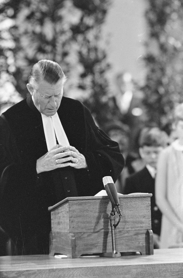 Ein Richter in einer schwarzen und weißen Robe spricht an einem Podium mit Mikrofon, umgeben von ein paar Menschen und Bäumen im Hintergrund.