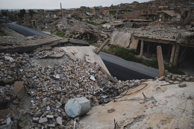 A destroyed building in Aleppo, Syria, with rubble and debris scattered across the street, a road in the foreground, and a sky in the background.