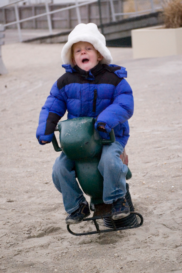 Ein kleiner Junge in einer blauen Daunenjacke sitzt auf einem Eisenfedersessel, mit einem weißen Rohrgeländer im Hintergrund.