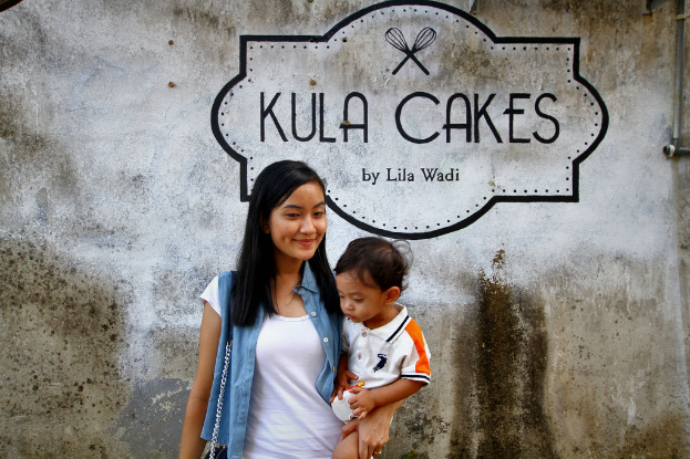 Eine Frau in einem weißen T-Shirt und blauer Jacke lächelt, waß ein Baby haltend, vor einer Wand mit Logo und Text.