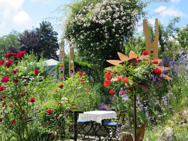 A garden with colorful flowers and plants, featuring a table with a cloth and chairs in the center, surrounded by trees under a cloudy sky at the Chelsea Flower Show.