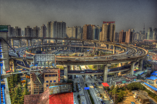 Stadtansicht mit einer zentralen Ringstraße mit Fahrzeugen, einer unteren Straße mit Autos und Fahrrädern, zahlreichen Hochhäusern im Hintergrund, einem Himmel oben, Bäumen auf der linken unteren Seite und kleinen Häusern überall in der Stadt verstreut.
