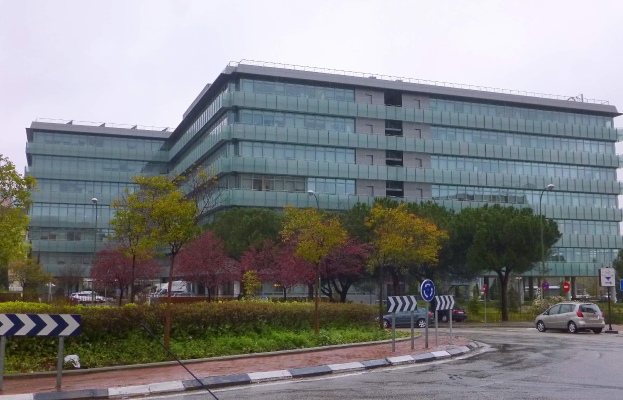 Außenansicht des Hauptsitzes der Europäischen Zentralbank, ein großes Gebäude mit vielen Fenstern, umgeben von Bäumen, Straßenlaternen und Fahrzeugen, unter einem bewölkten Himmel.