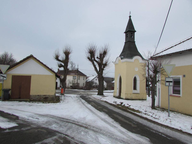 Ein kleines verschneites Dorf mit einer zentralen Kirche, Häusern mit Dächern und Fenstern, Schildern an Pfählen, einem Müllcontainer, einer Gruppe von Bäumen, Stromleitungen und einem bewölkten Himmel.