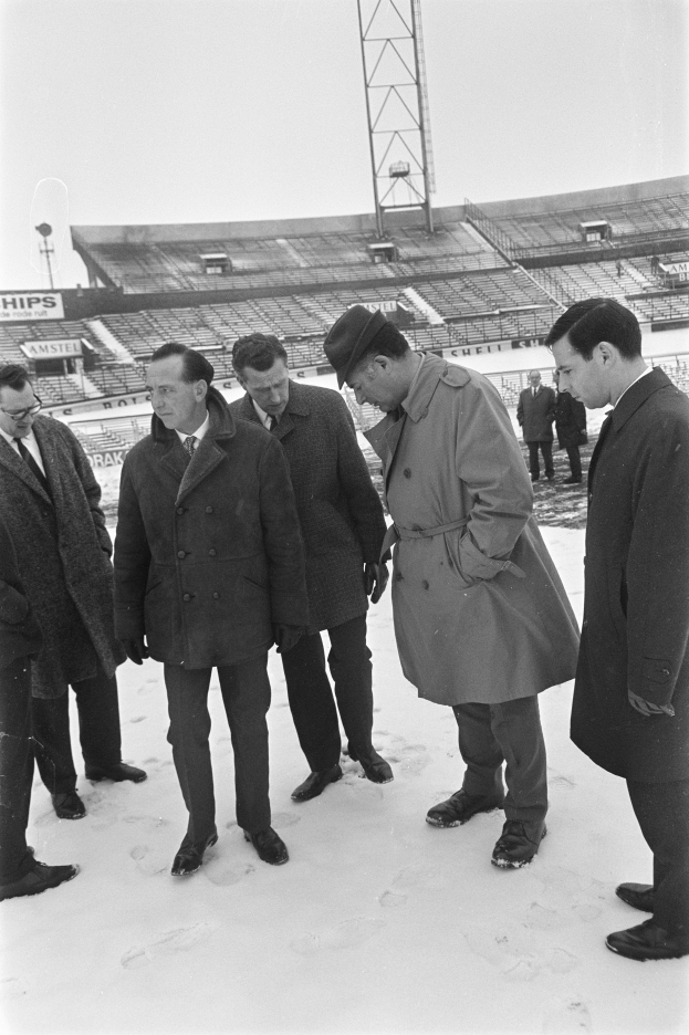 Eine Gruppe von Männern in Mänteln und Schuhen steht zusammen im Schnee, mit einem Stadion, einem Turm und einem klaren Himmel im Hintergrund.