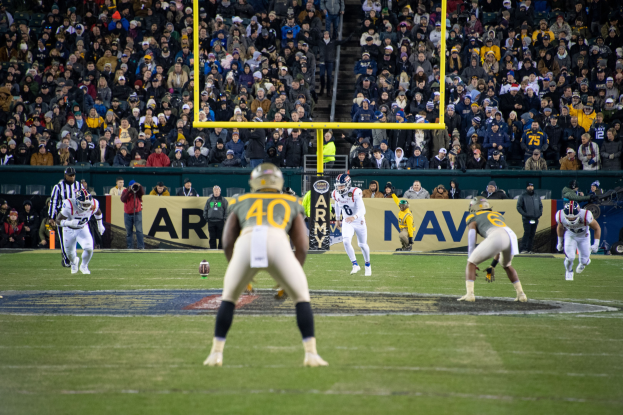 Fußballspieler in Sportkleidung und Helmen konkurrieren auf einem Feld mit Zuschauern im Hintergrund, Polen und Tafeln mit Text sichtbar.