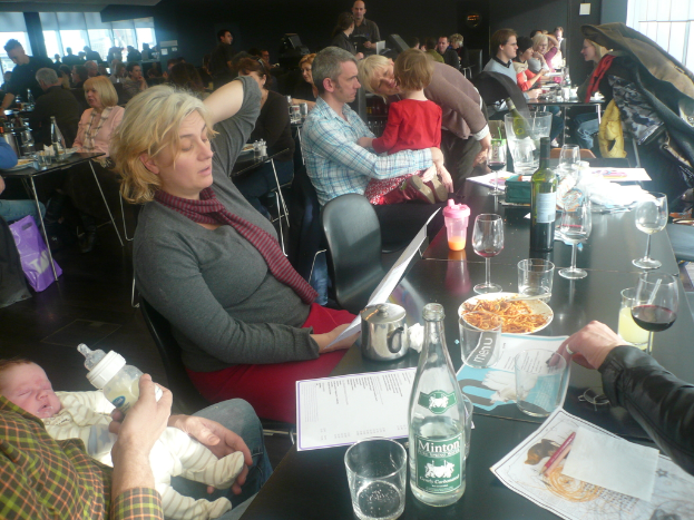 Ein Innenblick in ein belebtes Restaurant mit Gästen an Tischen, die mit Gläsern, Tellern mit Essen, Getränkeflaschen, Speisekarten und anderen Gegenständen gedeckt sind.