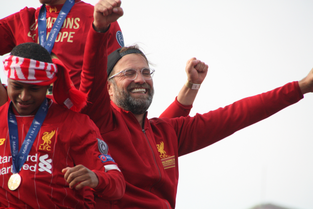 Liverpool-Trainer Jürgen Klopp feiert mit seinen Spielern den Sieg in der UEFA Champions League, umgeben von zwei medaillentragenden Personen und dem Himmel im Hintergrund.