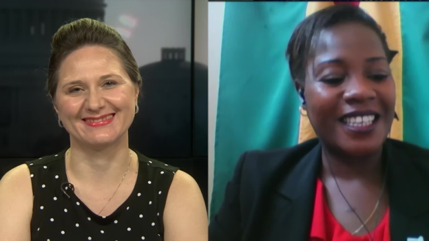 Zwei Frauen, die während eines Videoanrufs lächeln, eine in einem schwarzen und weißen Kleid, die andere in einer unbestimmten Farbe, mit einer Fahne, die an der Wand hinter ihnen in einer öffentlichen Einrichtung hängt.