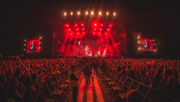 Eine große Menschenmenge steht vor einer hell erleuchteten Bühne mit Musikern, während andere von hinter den Absperrungen beobachten.