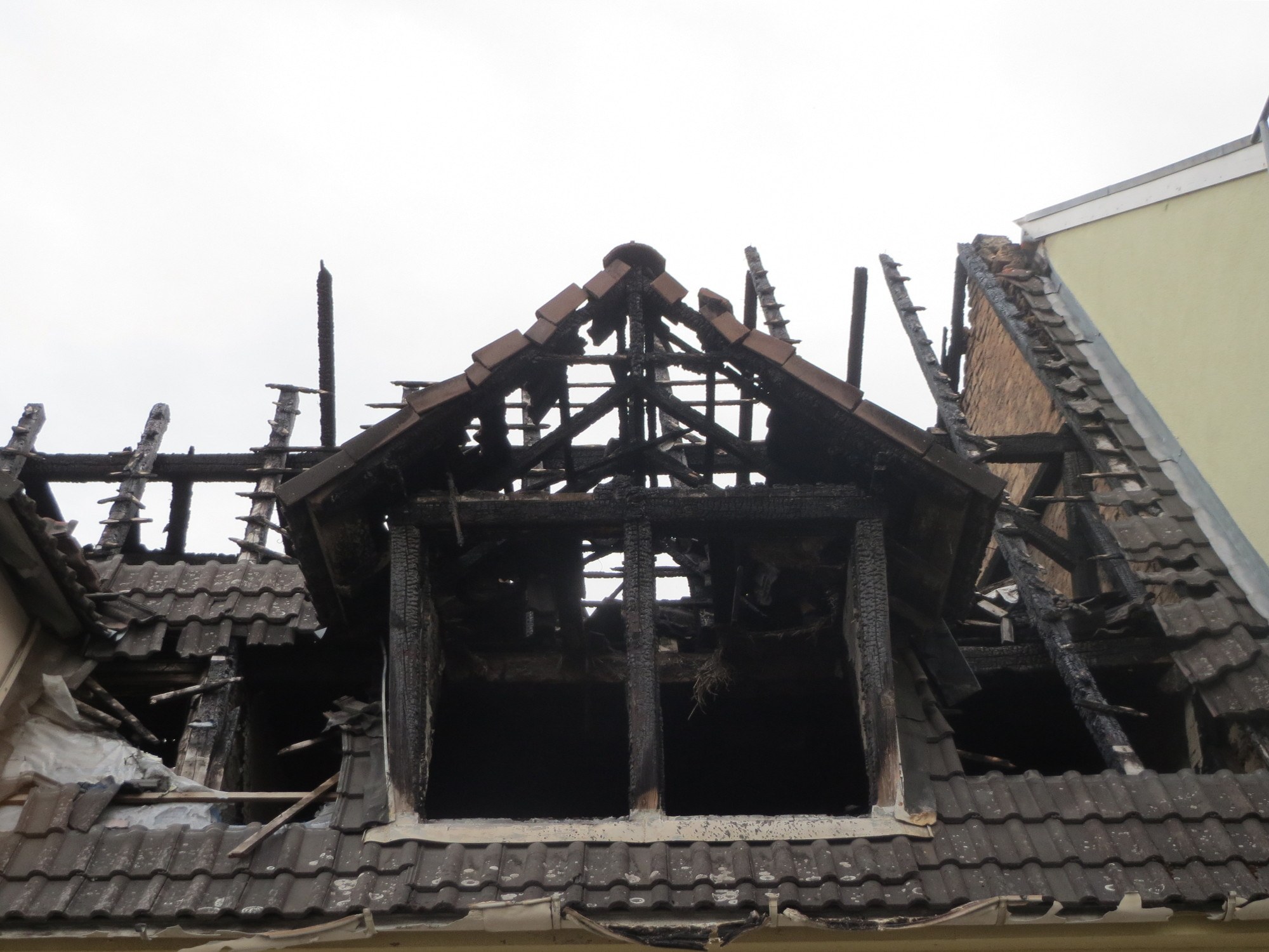 Brandgeschädigtes und einstürzendes Dach eines Gebäudes mit verstreuter Trümmer, vor einem Himmel-Hintergrund.