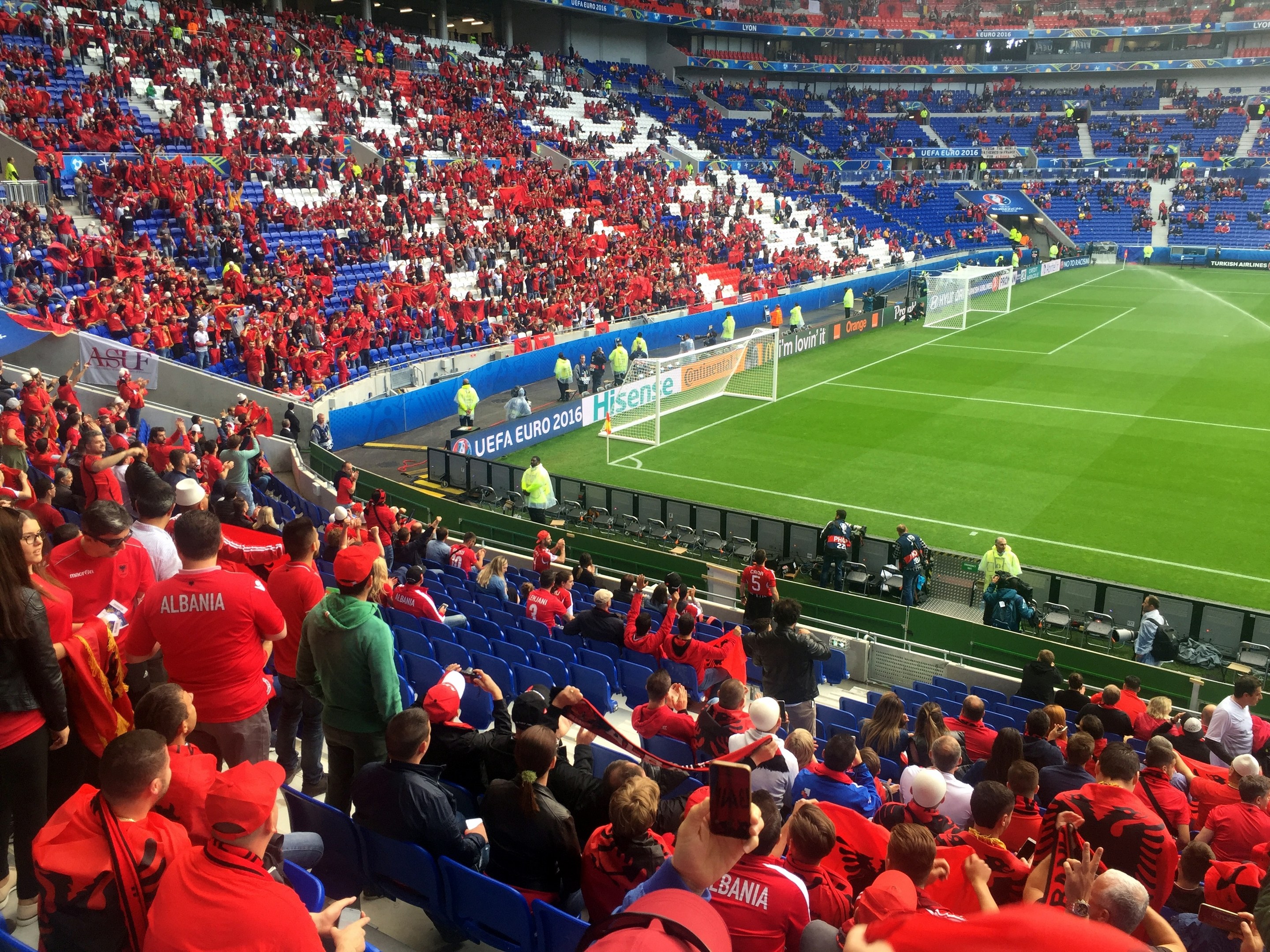 Eine große Menschenmenge in einem Stadion schaut einem Fussballspiel zu, mit Sitzplätzen und Stehplätzen, Fahnen, einem Torpfosten und Deckenbeleuchtung.