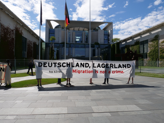 People holding a banner reading "Deutschland, Lagerland Migration is Not a Crime" in front of a building with windows, surrounded by grass, a metal fence, plants, trees, and flags with poles under a cloudy sky.