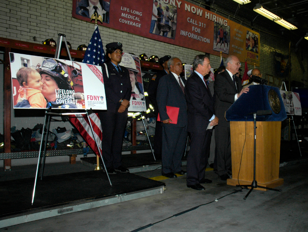 Eine Gruppe von Menschen steht in der Nähe eines Podiums mit Mikrofonen, umgeben von Schildern auf Ständern, Flaggen und Schildern an der Wand, alles unter hellen Lichtern.