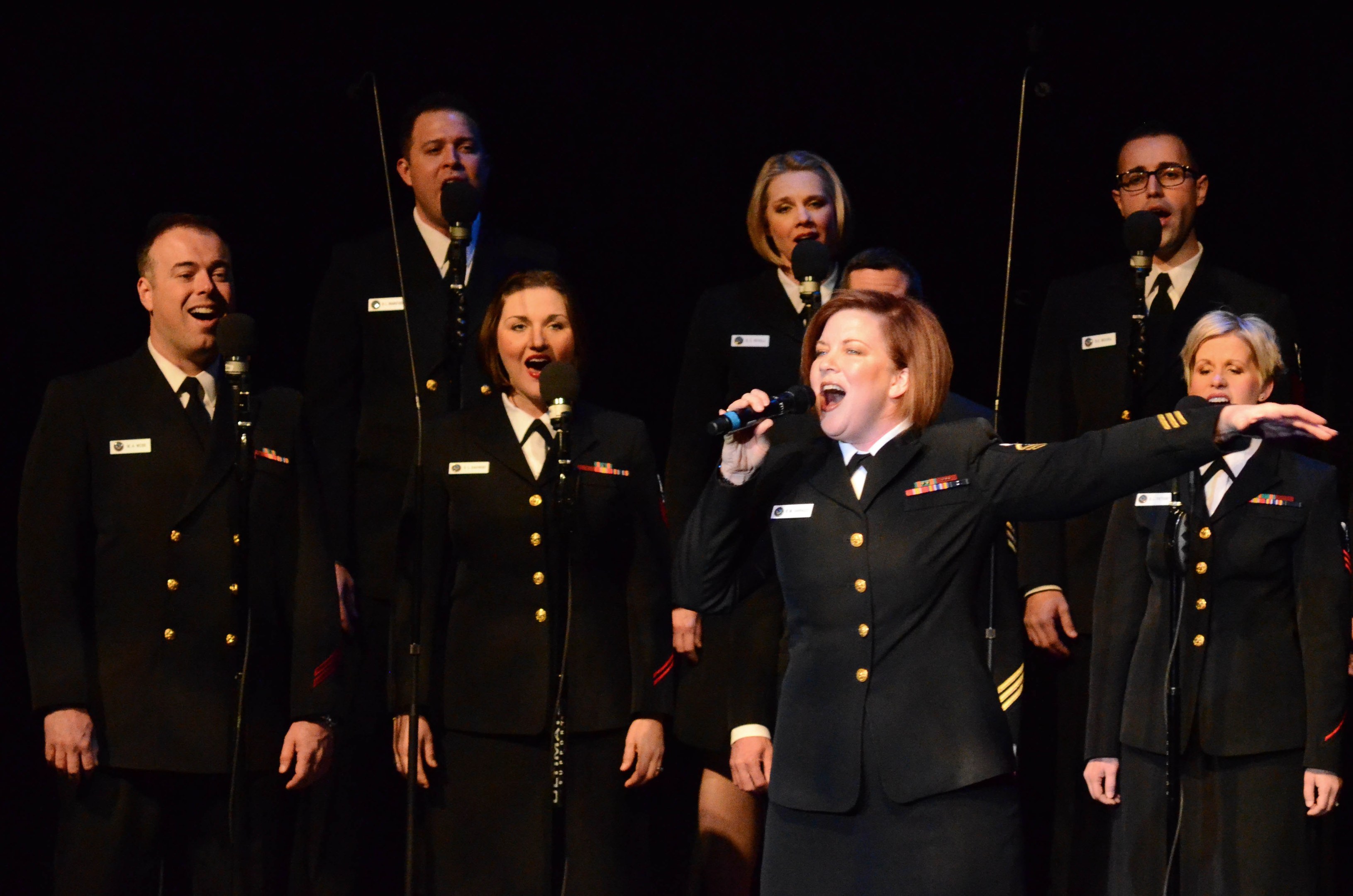 Group of people in military uniforms singing on stage with microphones, dark background.