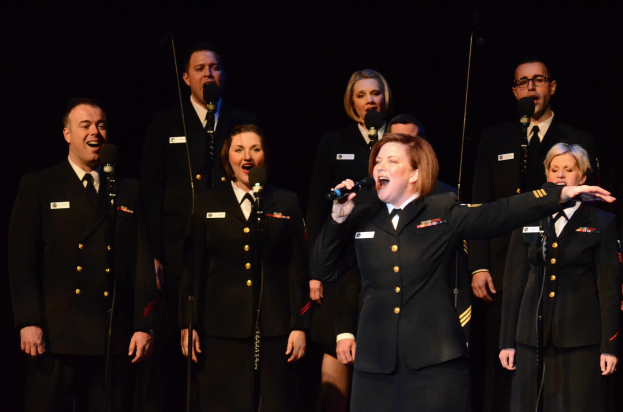 Group of people in military uniforms singing on stage with microphones, dark background.