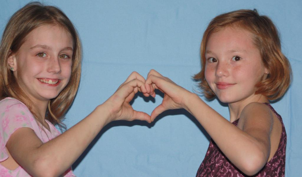 Zwei Kinder lächeln und bilden mit den Händen ein Herz vor einer Wand.