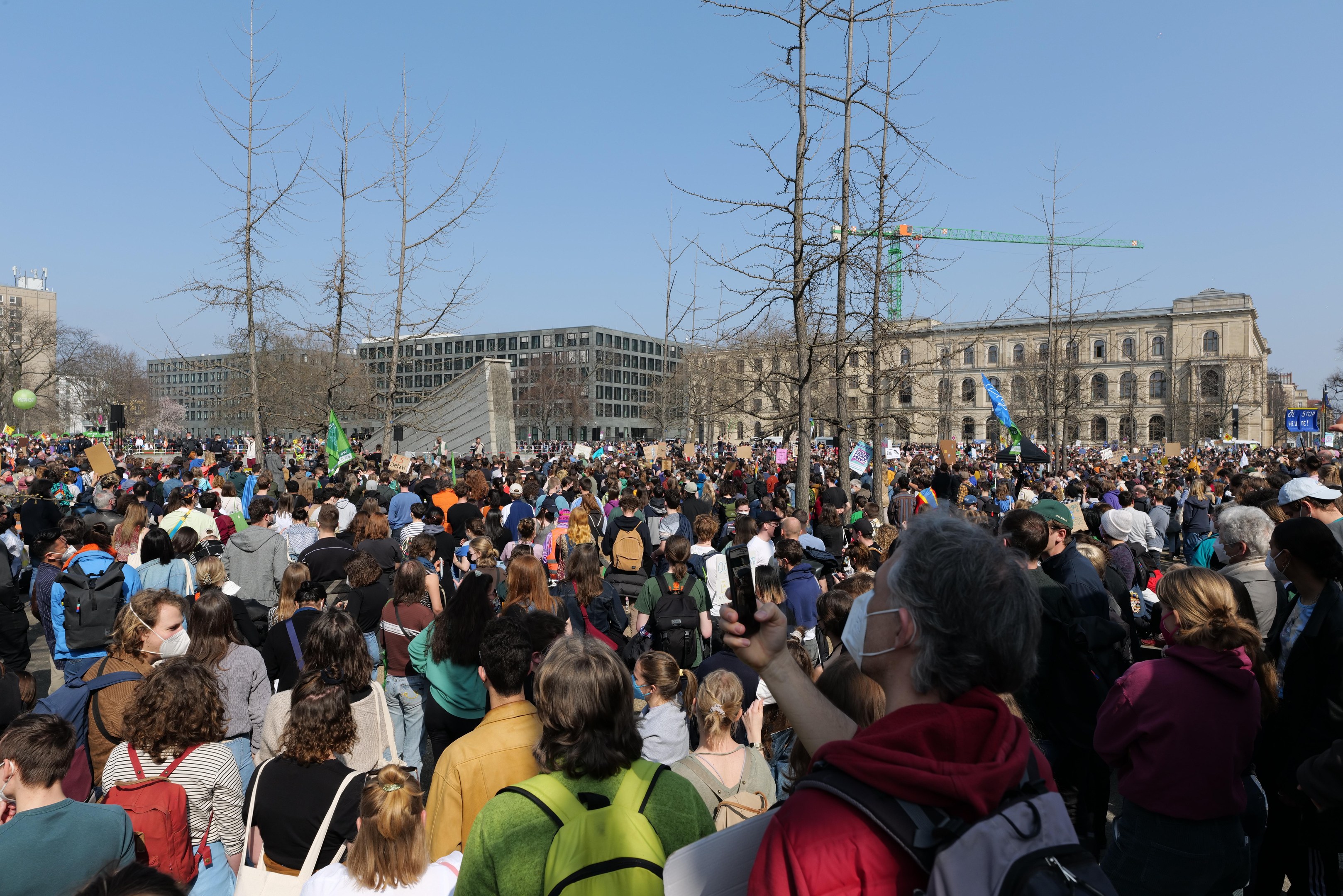 A large crowd of people participating in a climate change protest, standing in front of a building with trees and a clear sky in the background, many holding placards and wearing bags.
