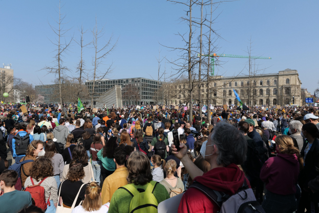 A large crowd of people participating in a climate change protest, standing in front of a building with trees and a clear sky in the background, many holding placards and wearing bags.