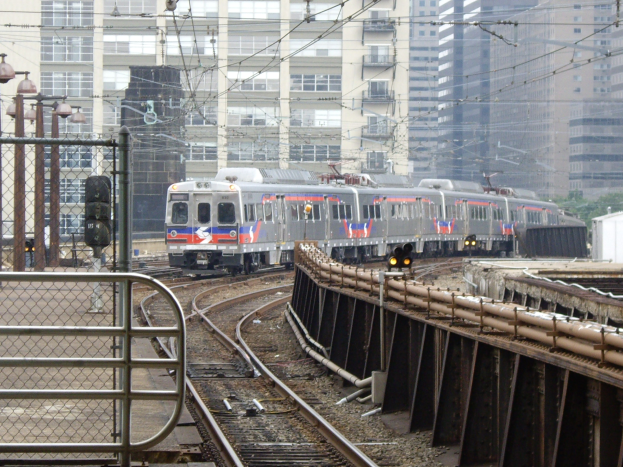 A commuter train travels on tracks beside tall buildings with various urban infrastructure elements visible in the background.