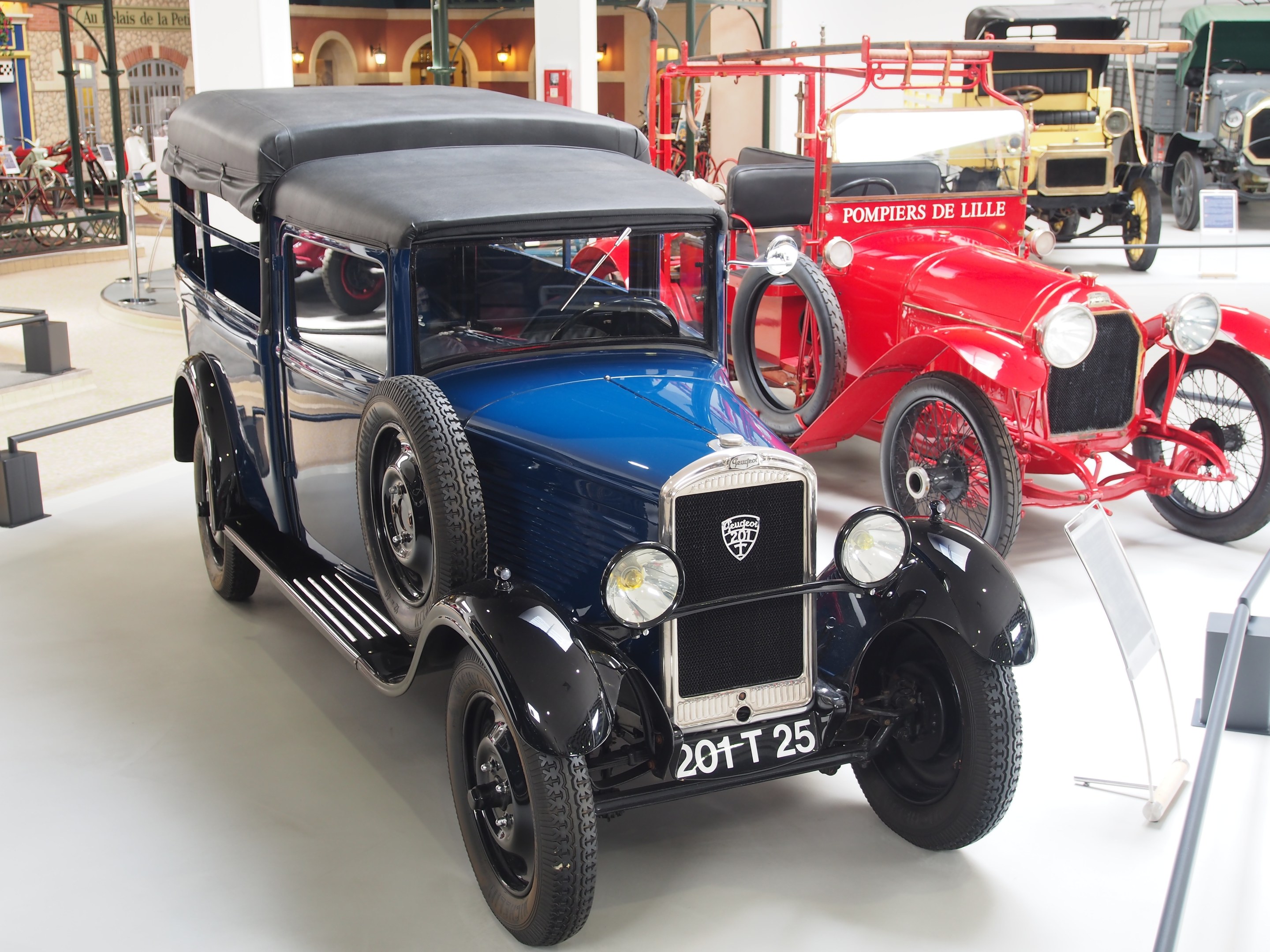 A vintage car displayed in a museum among other classic vehicles, with roped stands to the left and informational boards and pillars in the background.