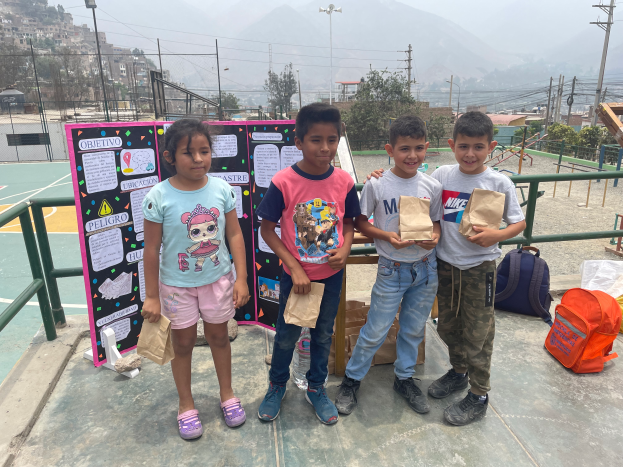 Eine Gruppe von Kindern steht auf einem Basketballfeld, jedes hält eine Papiertüte, mit Vorräten und einer Texttafel in der Nähe und einer Kulisse aus Zäunen, Pfosten, Bäumen, Gebäuden und Hügeln.