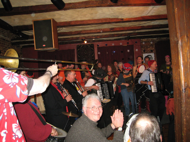 Das Innere eines Gebäudes mit sitzenden und stehenden Menschen, drei Vorhänge, ein Mikrofon auf einem Ständer, Musikinstrumente, Sterne, ein Lautsprecher, Kabel und eine Deckenleuchte.