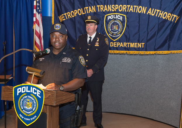 Zwei Polizisten stehen vor einem Rednerpult, einer hält Papiere und spricht in ein Mikrofon, mit einer Fahne, einem Tisch und einer Tafel mit Symbolen und Text im Hintergrund.