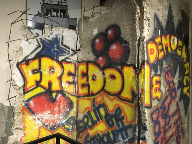 Mauer mit Graffiti, das "Freiheit und Demokratie" in großen schwarzen Buchstaben liest, unter einem Geländer und über einem Gebäude mit Fenstern im Hintergrund.