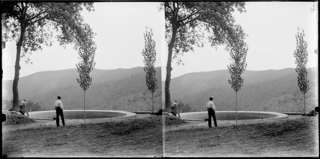 Schwarzes und weißes Foto eines Mannes, der vor einem Teich mit Bäumen, Hügeln und einem klaren Himmel steht.