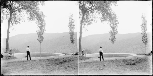 Schwarzes und weißes Foto eines Mannes, der vor einem Teich mit Bäumen, Hügeln und einem klaren Himmel steht.
