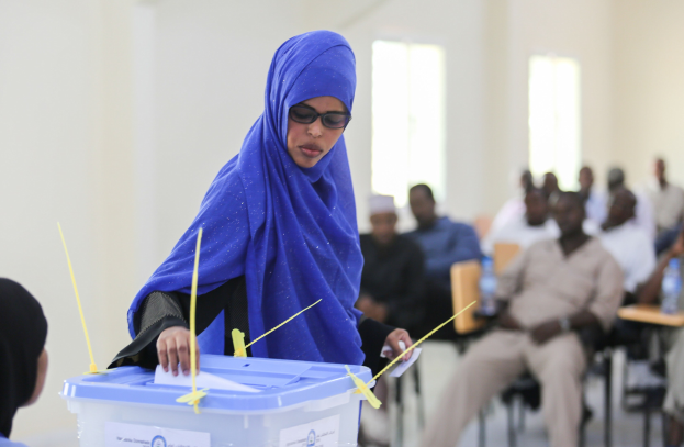 Eine Frau mit blauem Kopftuch stimmt in einer Wahlkabine ab, während andere in einem Raum mit Fenstern in der Nähe sitzen.