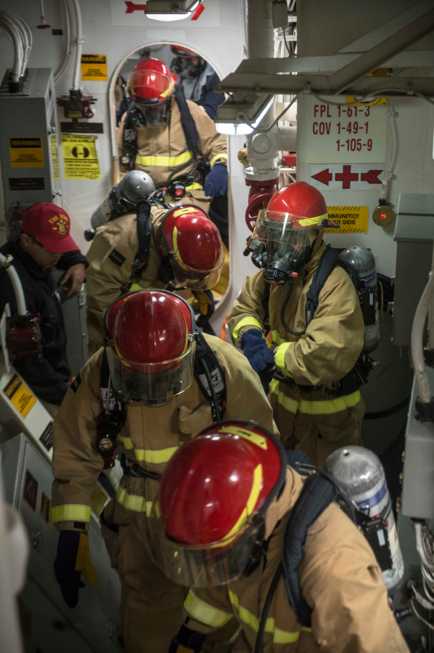 Eine Gruppe von Feuerwehrleuten in Schutzausrüstung, einschließlich Helmen, Handschuhen und Sauerstoffflaschen, die an einem Boot mit Rohren und anderen Gegenständen im Hintergrund arbeiten und Plakate mit Text an der Wand.
