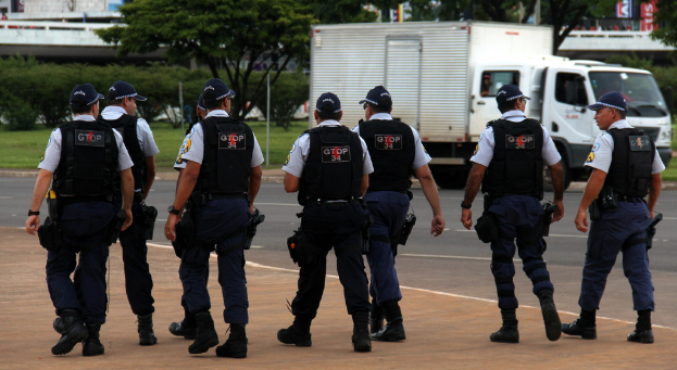 Eine Gruppe von Beamten in Uniform, einschließlich Mützen, Hemden, Schutzwesten, Uhren, Hosen und Schuhen, die auf einer Straße mit einem geparkten Lkw auf der rechten Seite neben einem Zaun, Gebäuden, Bäumen, Pflanzen, Polen und Bannern im Hintergrund und einer Brücke im oberen rechten Eck gehen.
