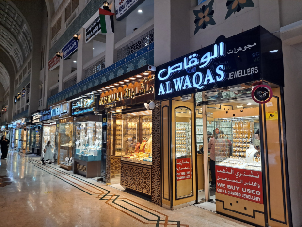 A bustling shopping mall in Dubai, UAE, featuring people walking, stores with name boards, bright lights, flags, and a variety of jewelry displays.