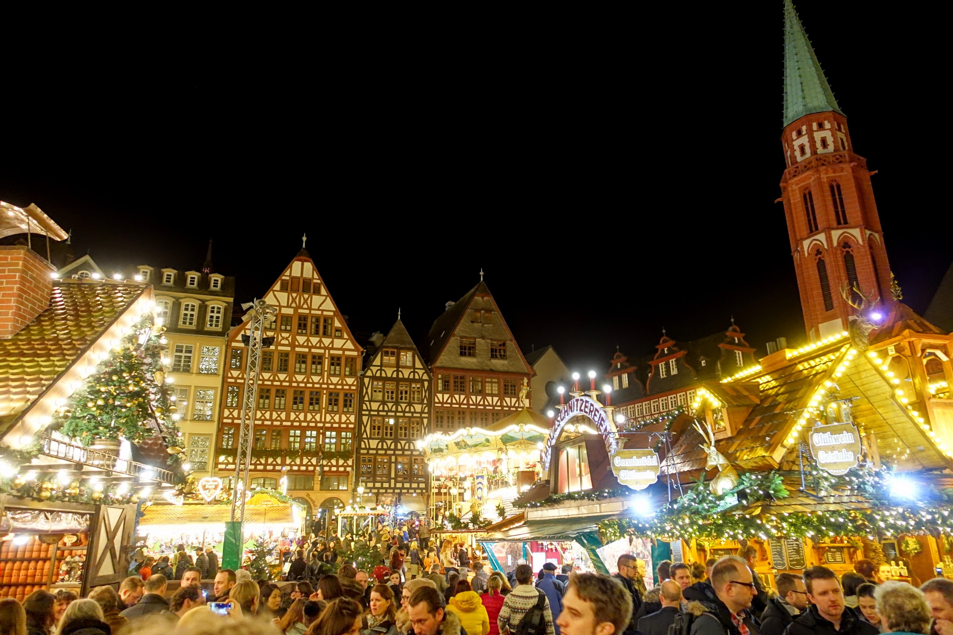 Ein geschäftiges Weihnachtsmarkt in Nürnberg, Deutschland mit Menschen um geschmückte Stände, festliche Lichter, Gebäude mit Fenstern, ein Uhrenturm und einen dunklen Himmel.