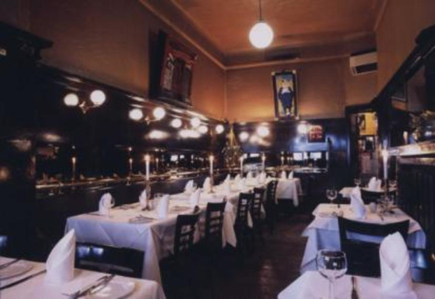 Ein Tisch mit Gläsern, Kerzen in Ständern, Servietten und Tellern, umgeben von Stühlen, mit Lampen und Rahmen an den Wänden.