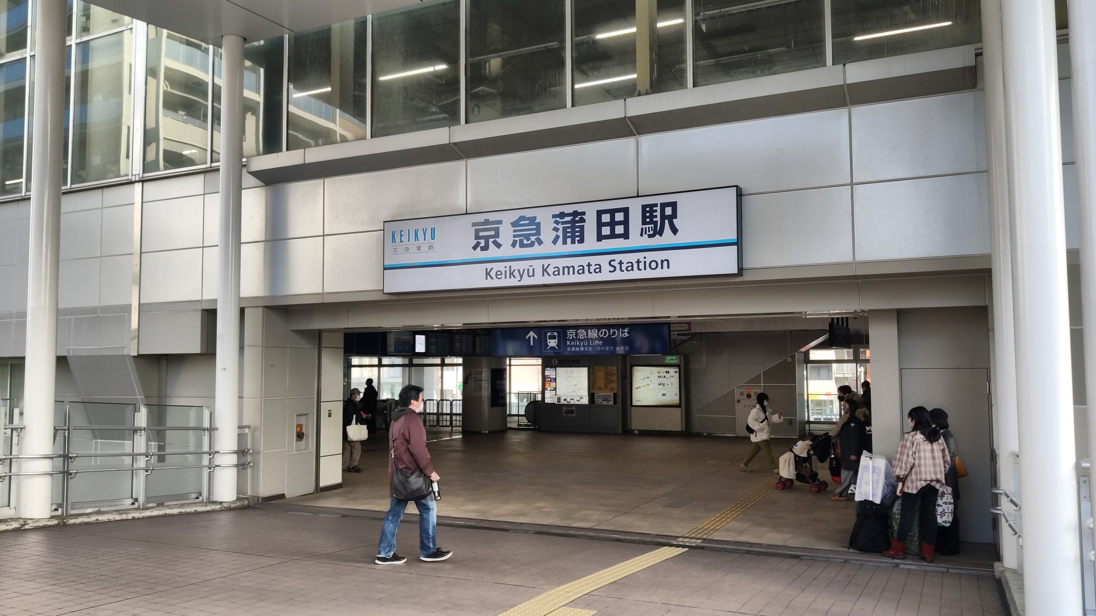 Menschen mit Taschen durch eine Bahnhofshalle gehen, ein Schild mit Text, Säulen, Geländer, Glaswände und Deckenlampen; ein Mann in Schuhen ist mittig sichtbar.