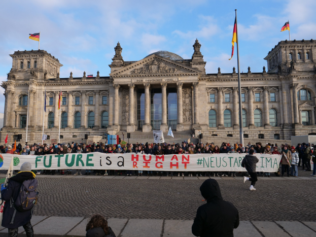 Group of people holding a banner with the text "Future is a Human Neustar ima" in front of the Reichstag building in Berlin, Germany, with its architectural details and surrounding flags visible under a cloudy sky.