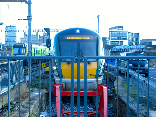 Zwei Züge auf der Schiene mit einem Zaun im Vordergrund, Masten auf dem Land, einer mit einer Signalbeleuchtung, ein Auto auf der Straße rechts, Gebäude im Hintergrund und der Himmel oben.