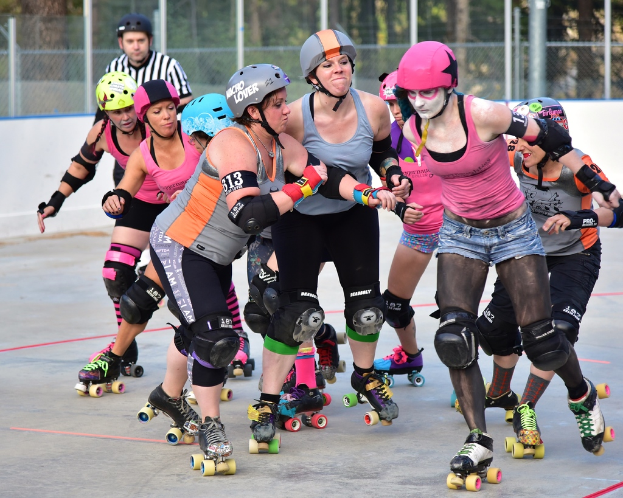 Mehrere Frauen mit Helmen und Skateschuhen schieben sich gegenseitig auf Skates, während ein Mann danebensteht, ein Metallzaun und B√§ume im Hintergrund zu sehen sind.