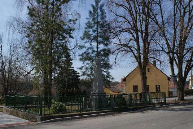 Ein Holocaust-Denkmal steht in der Mitte einer Straße, umgeben von einem Metallzaun, umgeben von Grün, Häusern und einem bewölkten Himmel.